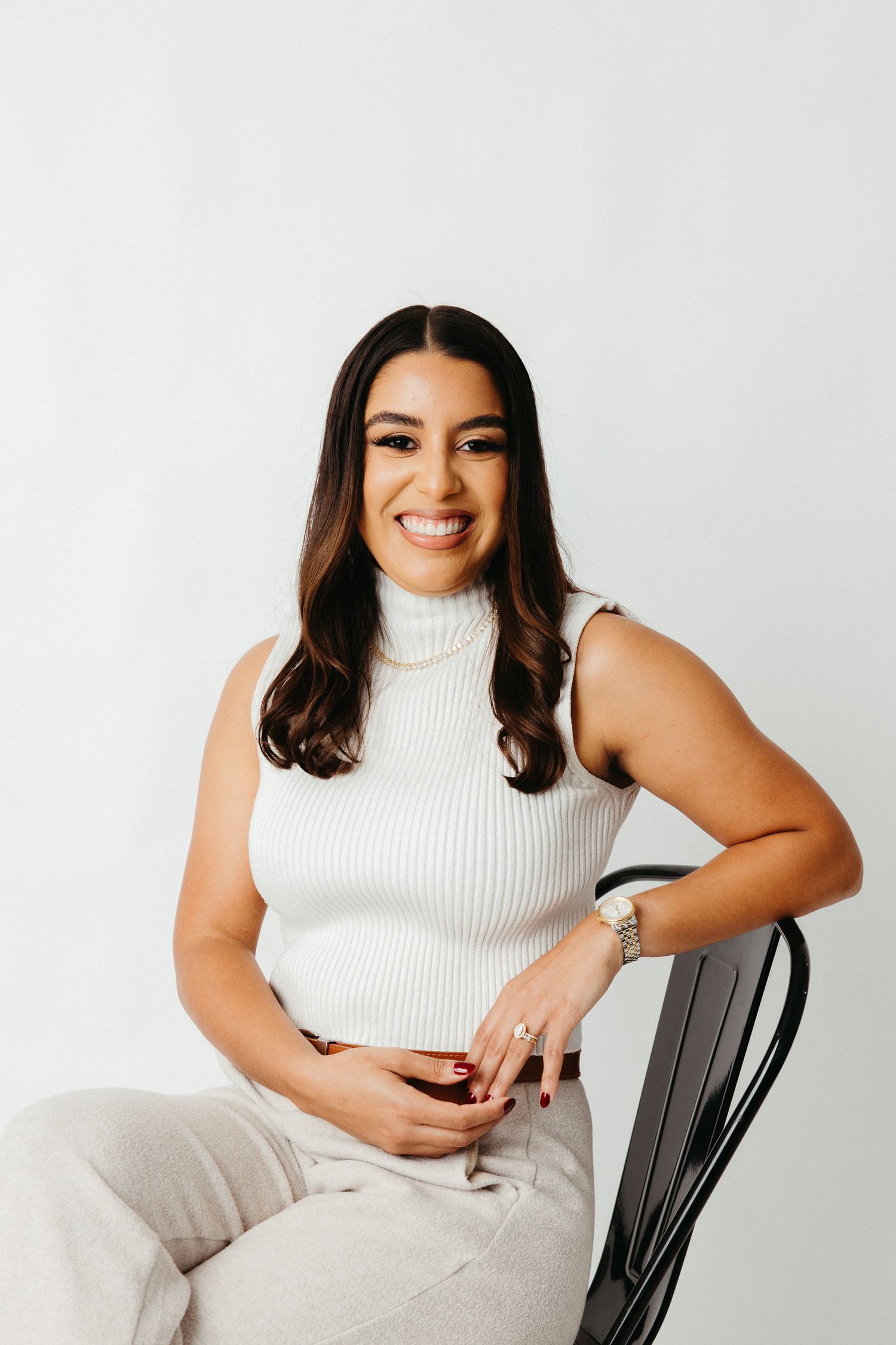 Portrait of a woman in white top and light pants sitting on a black chair against white background.