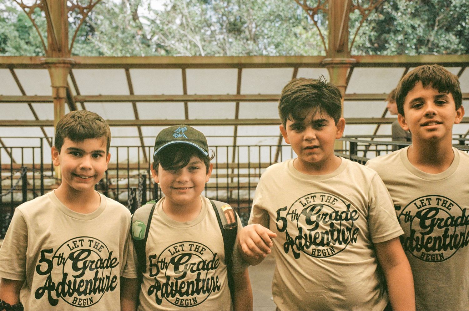 Four youths wearing matching beige t-shirts pose together at an outdoor venue.