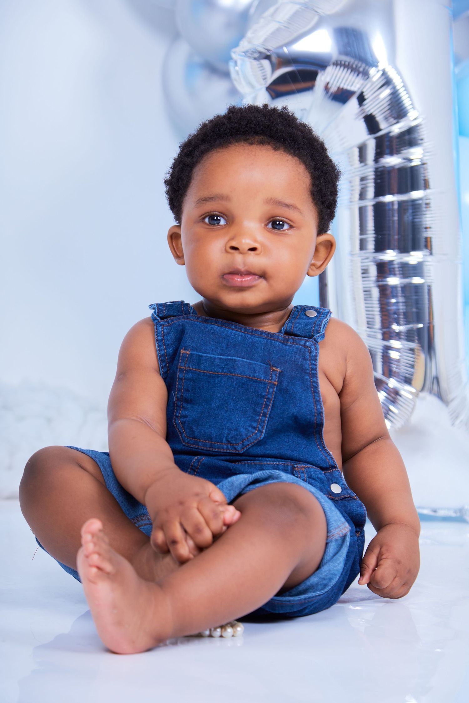 A baby in denim overalls plays on a white floor with silver decorations in the background.