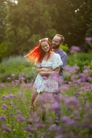 A couple embraces in a garden with blooming purple wildflowers as her red hair flows in the sunlight.
