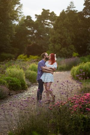 A couple shares a romantic moment amidst blooming purple wildflowers and greenery in a garden setting during sunset.