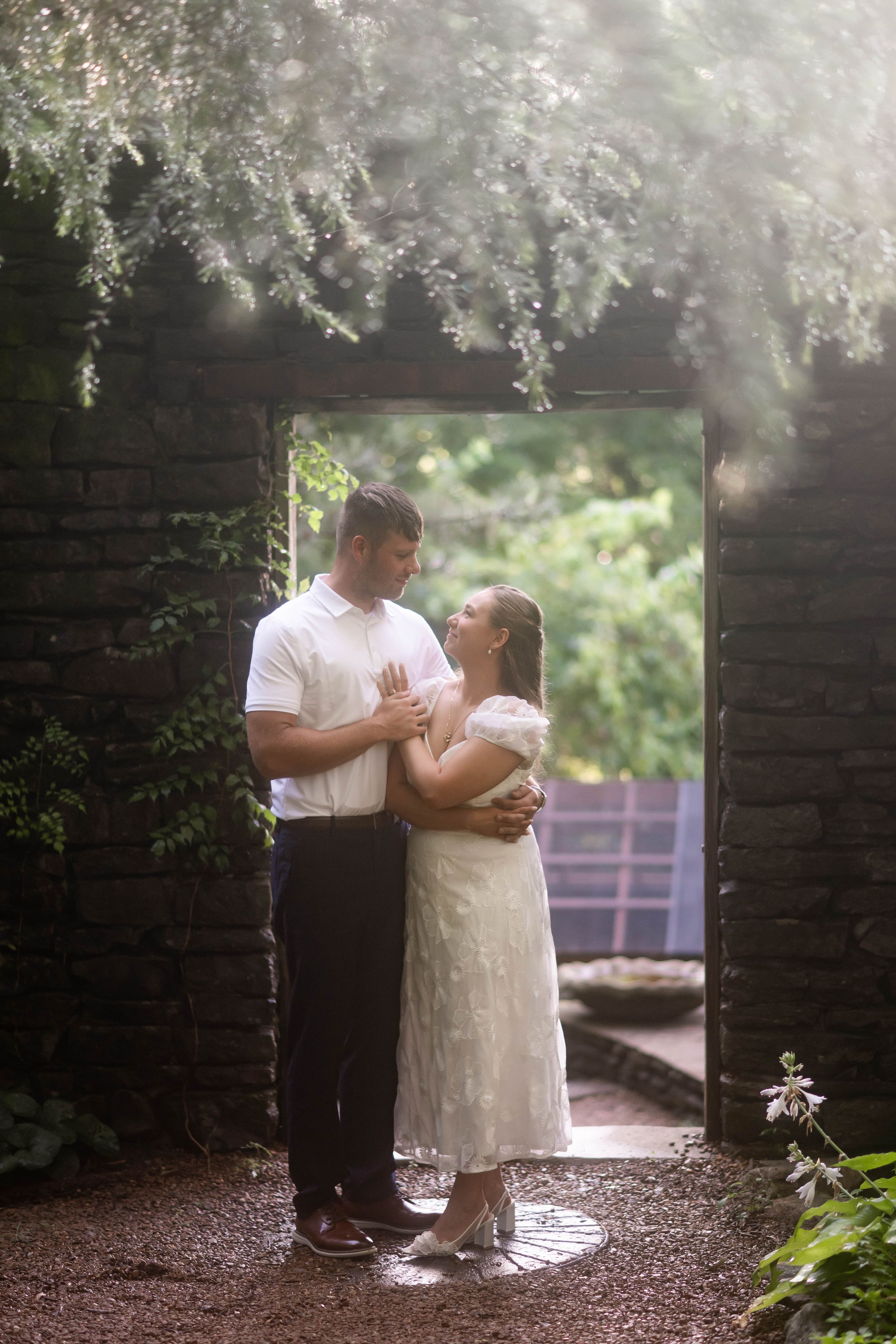 A romantic couple embraces in a garden archway framed by soft white flowers and greenery on a misty day.