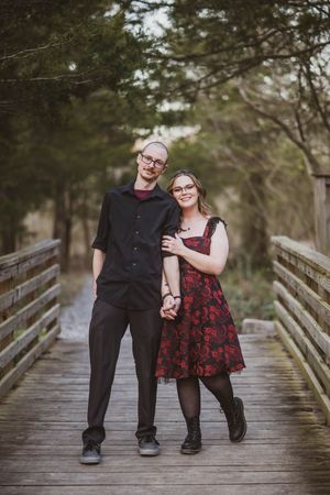 A couple stands together on a wooden boardwalk surrounded by trees, wearing coordinated formal attire.