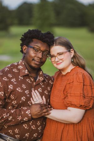 A couple wearing glasses and coordinating brown and orange outfits embrace in an outdoor setting with greenery.