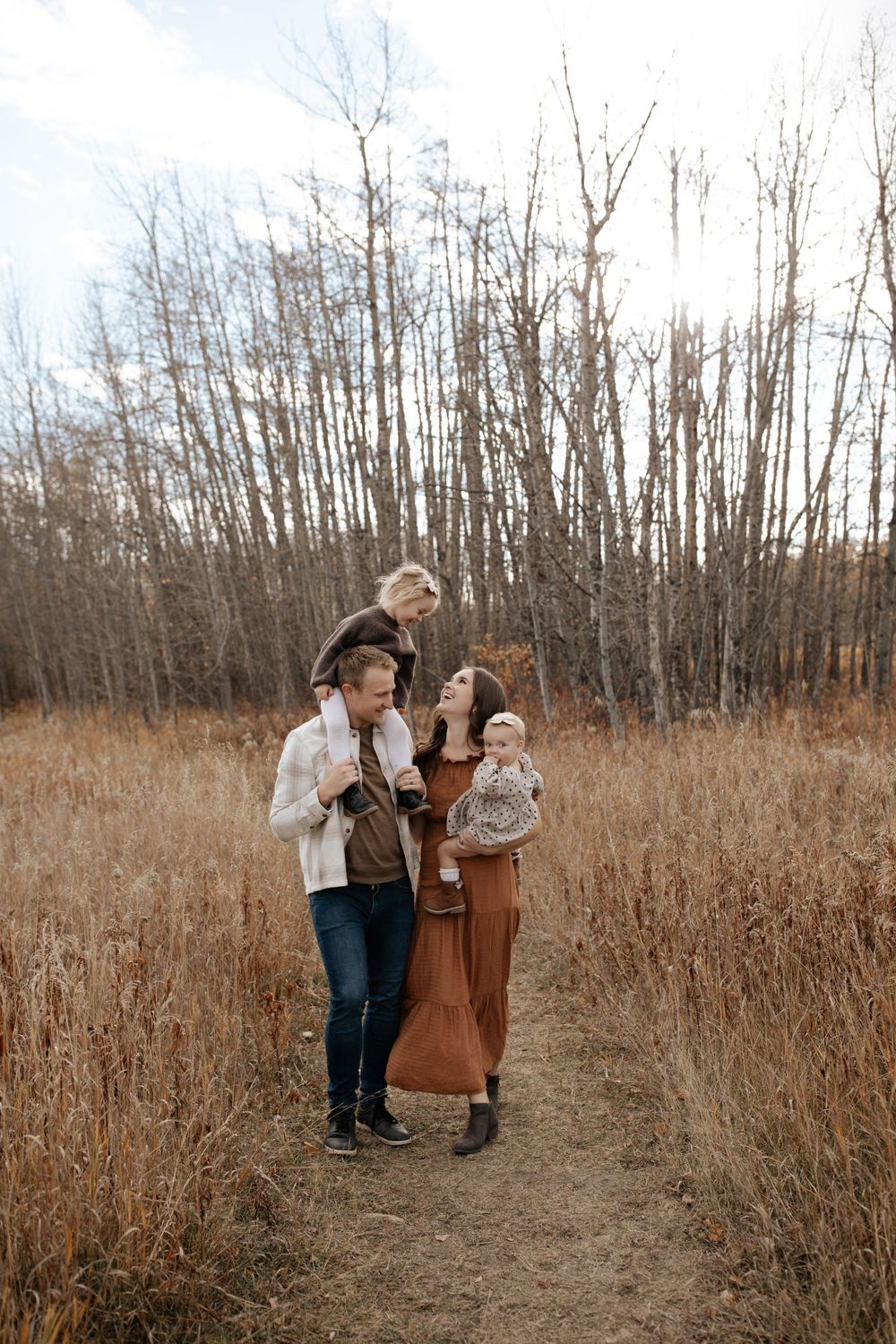 A family shares tender moments together in a golden autumn field with bare trees in the background.