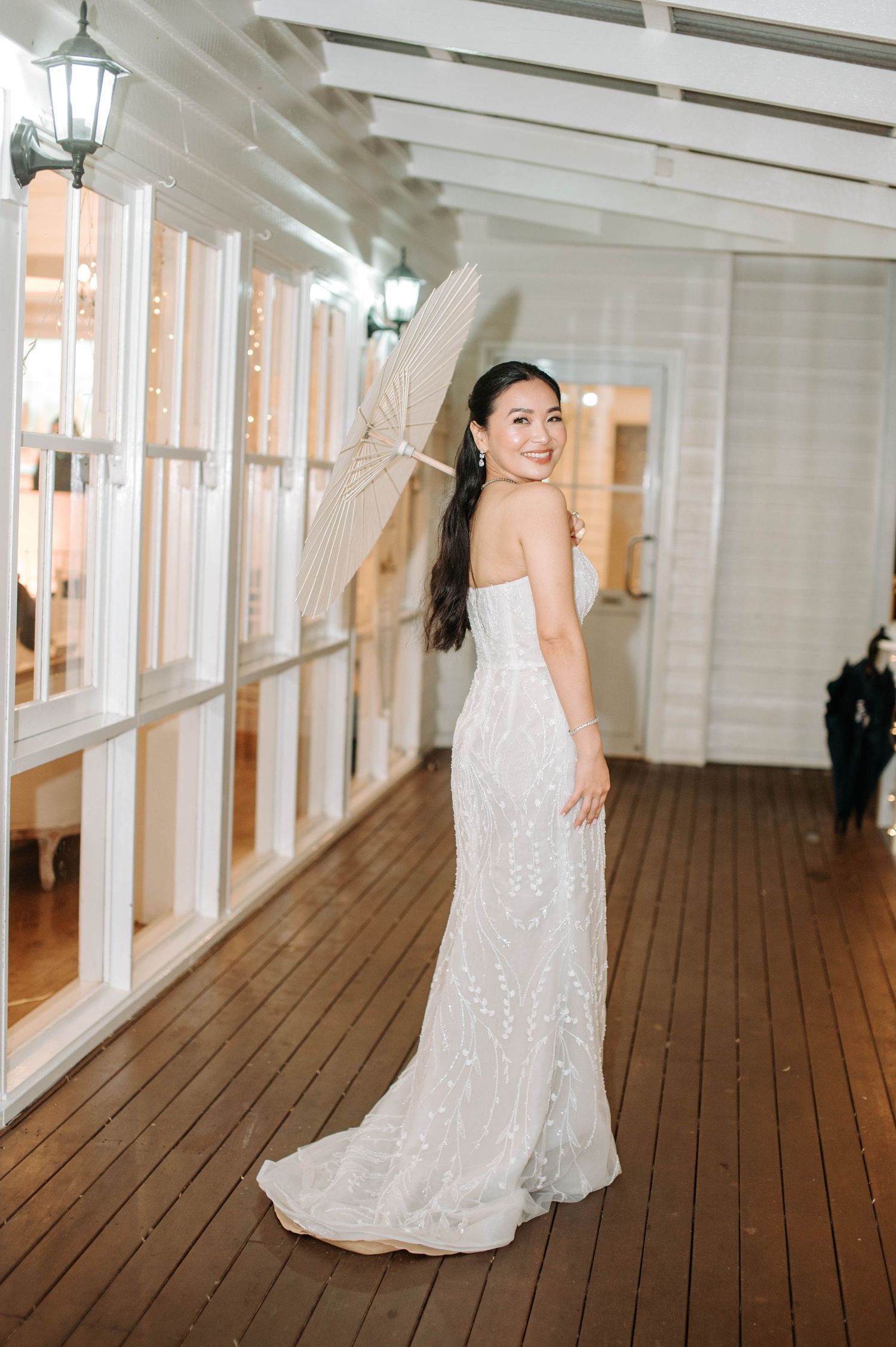 Bride in a fitted lace wedding gown poses in an elegant hallway with large windows and wooden floors.