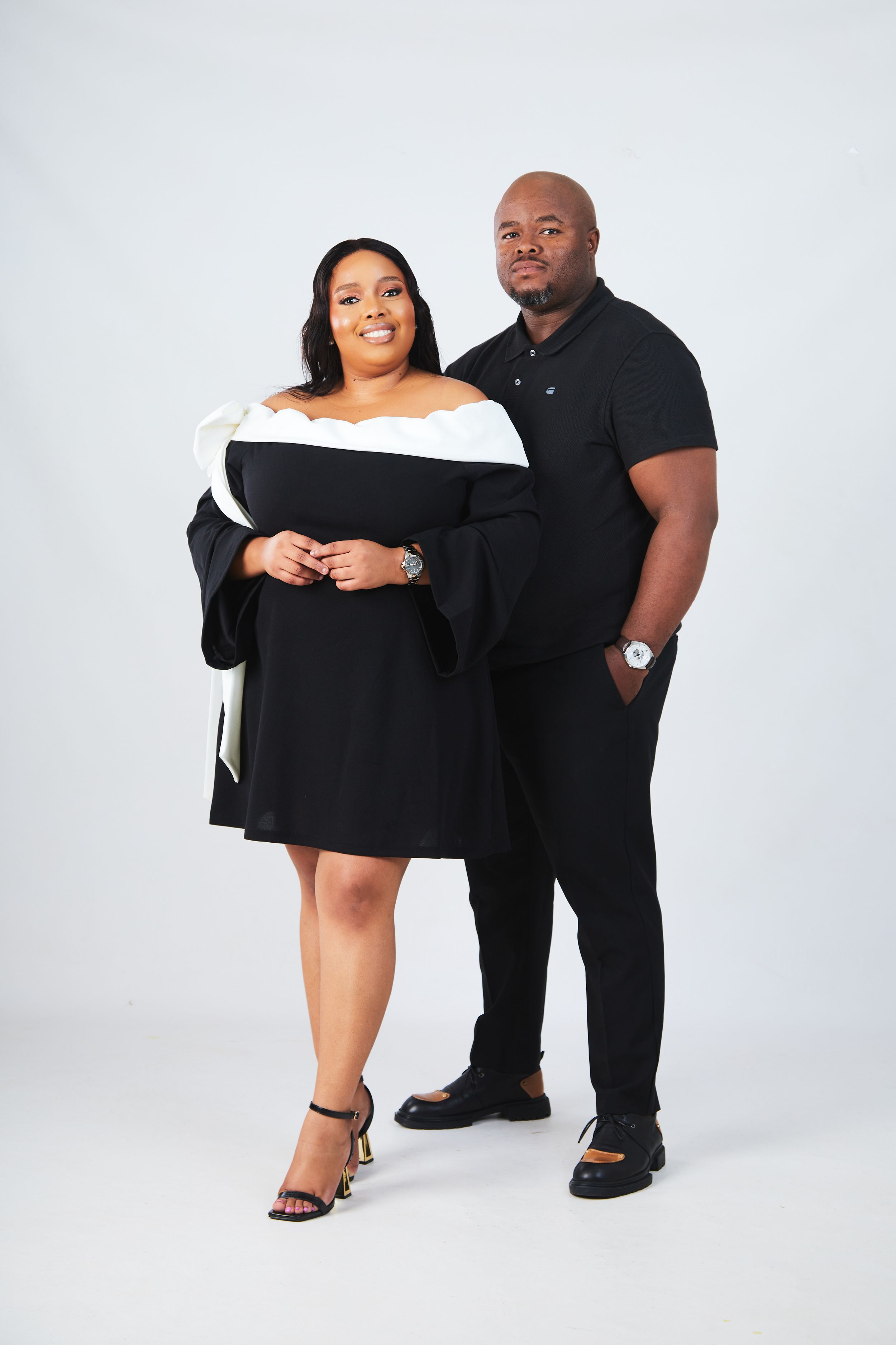 A couple poses together in matching black outfits for a stylish photo shoot against a white backdrop.