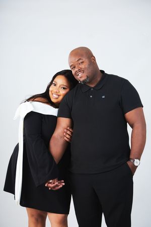 A couple wearing black outfits poses together against a white background in a studio portrait.
