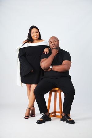 A couple poses together in a stylish black and white studio photoshoot on a wooden stool against a white backdrop.