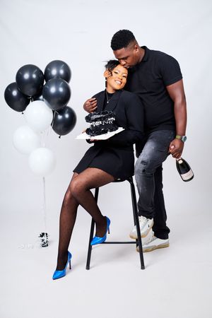 A couple in black attire poses playfully with balloons and champagne during a photo shoot.