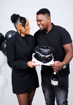 A couple in black outfits shares a fun moment with cake during a celebratory photoshoot.