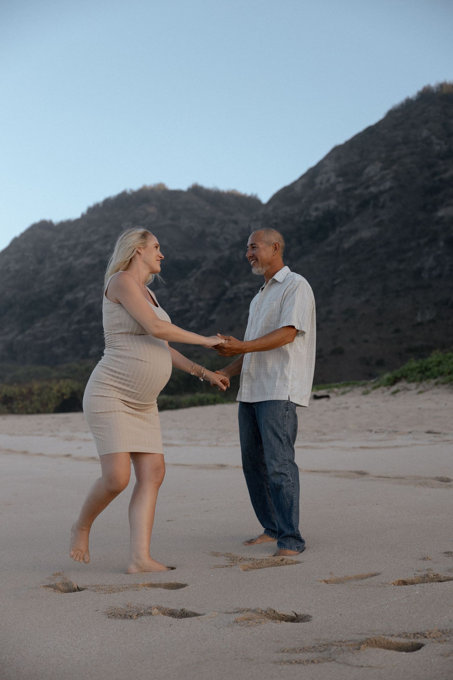A couple dancing together on a beach with mountains in the background, celebrating their pregnancy with a maternity photoshoot.