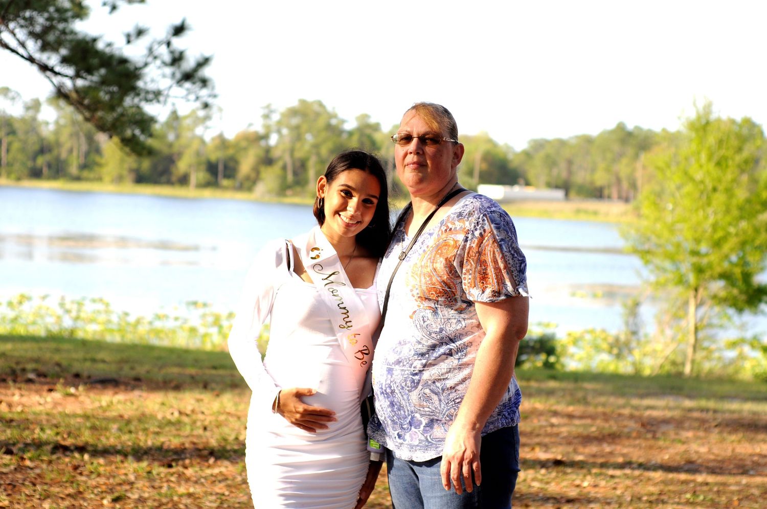 A couple stands together near a scenic lake at sunset, with trees and natural scenery in the background.