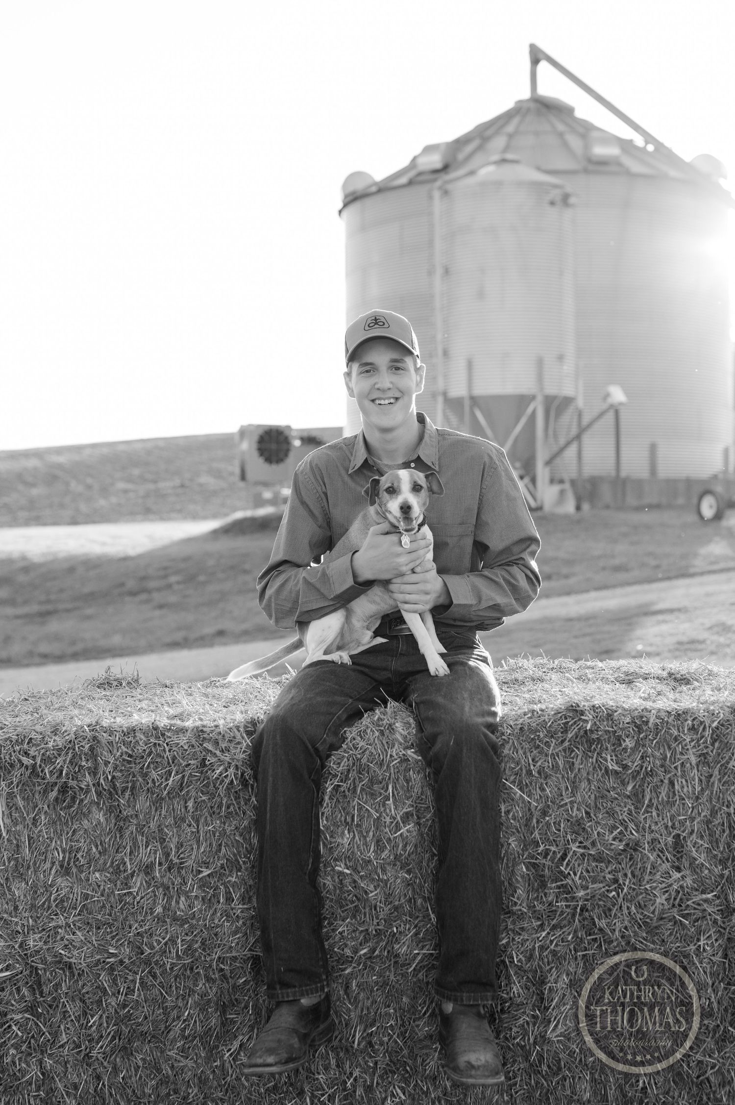 Black and white portrait of person with small dog sitting on hay bale by grain silo.