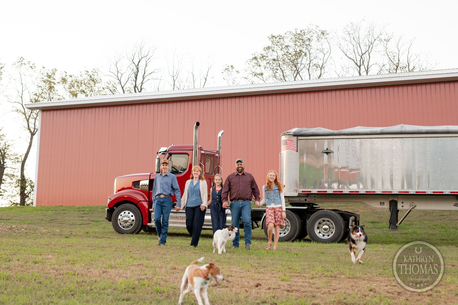 A group is gathered near a red truck with dogs playing in a grassy field by a farm building.