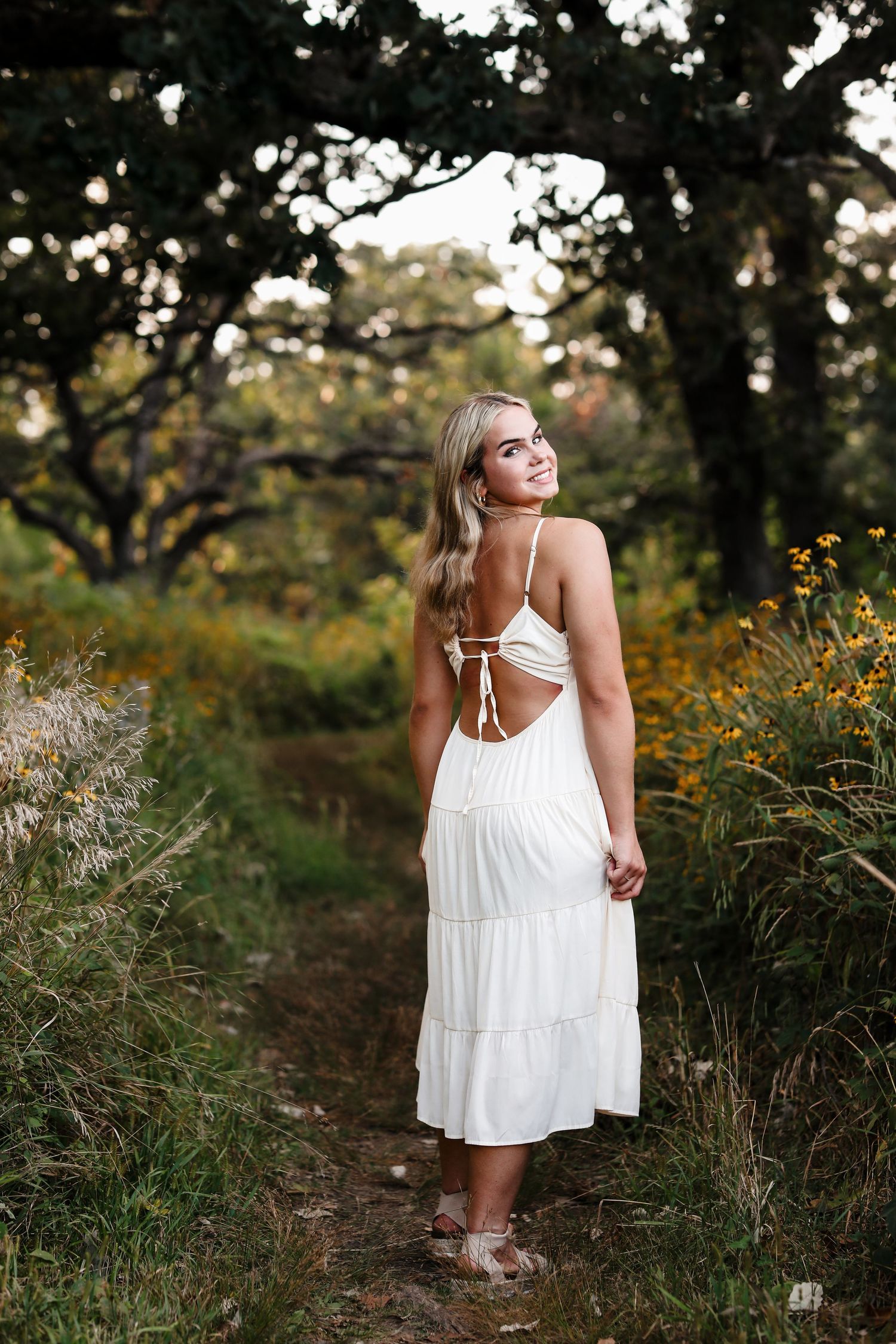 Blonde woman in white backless sundress standing on nature path surrounded by wildflowers.