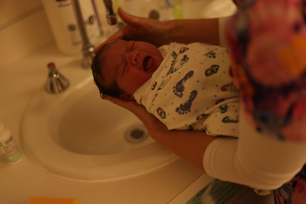 A newborn baby is gently supported over a bathroom sink during their first bath.