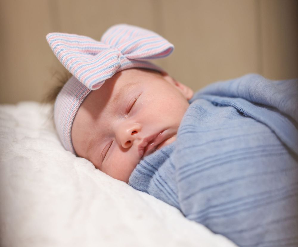 Peaceful newborn baby sleeping swaddled in blue with a pink striped bow hospital hat.