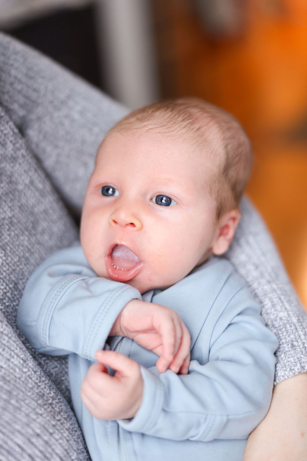 Alert blue-eyed baby boy in light blue onesie with tongue out sitting in a gray bouncer.