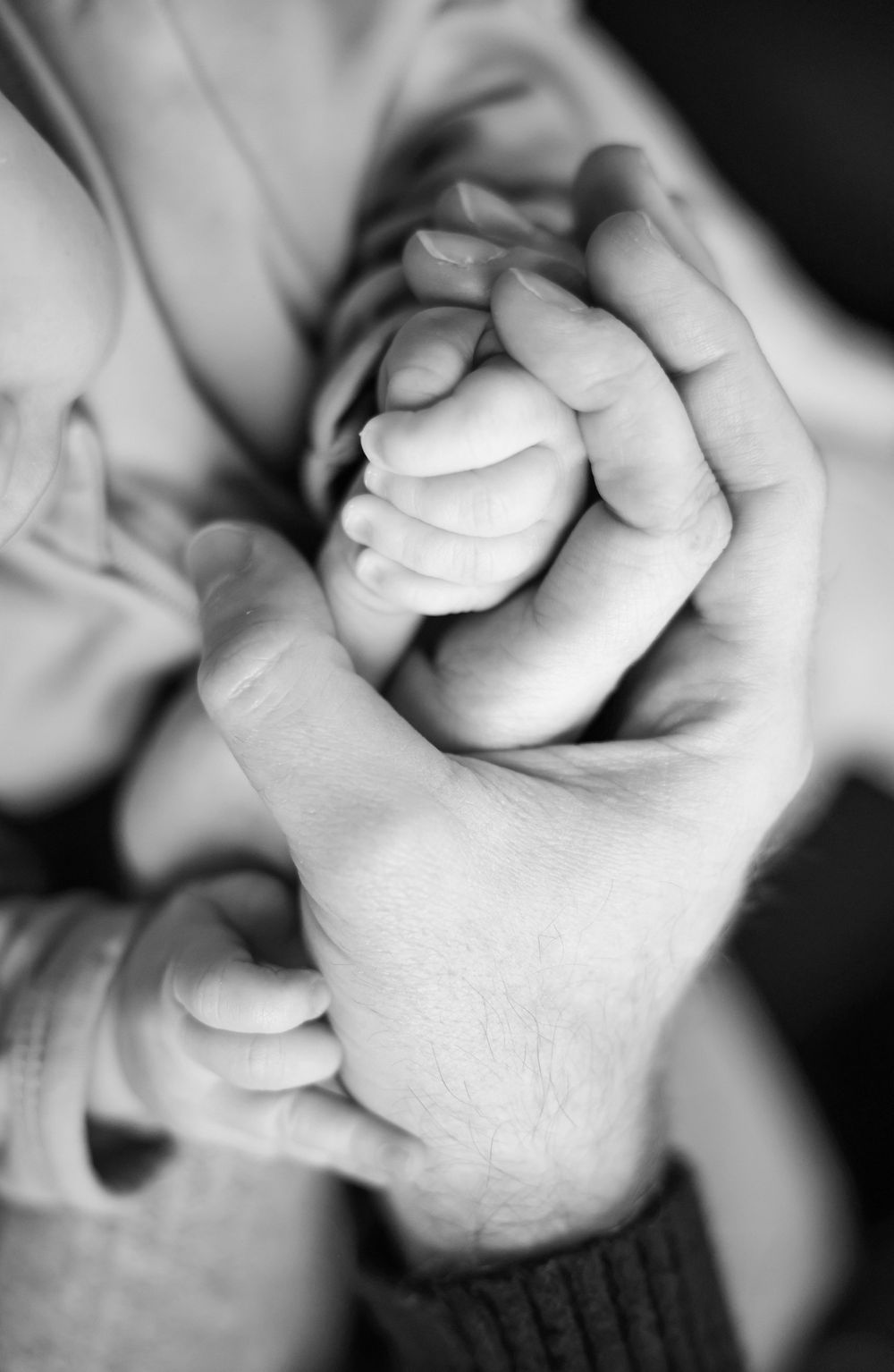 Black and white close-up of parents' hands gently cradling their newborn baby's tiny fist.