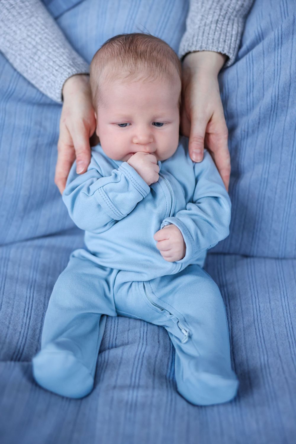 Newborn baby in blue sleeper held by mother's hands on blue sofa.