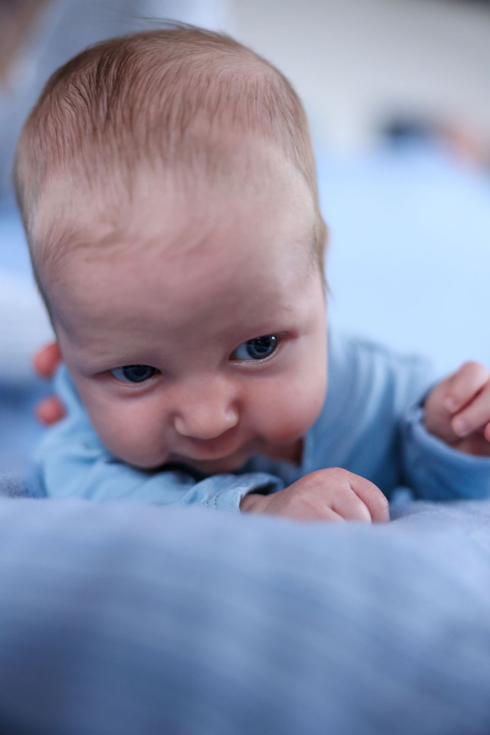 Newborn baby in blue outfit lifting head during tummy time on bed.