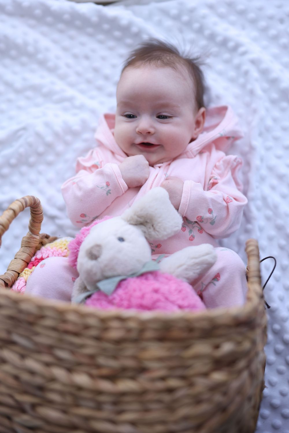 A cute baby girl in pink outfit lies smiling in a wicker basket with a stuffed bunny toy.