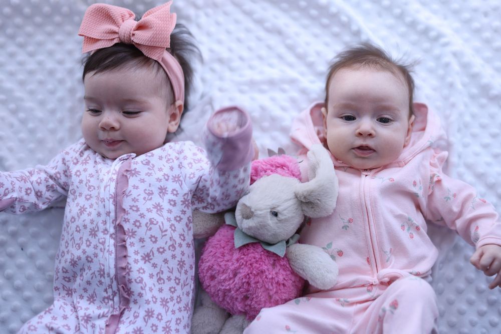 Two baby girls in pink outfits lying next to a stuffed bunny toy, one wearing a pink bow headband.
