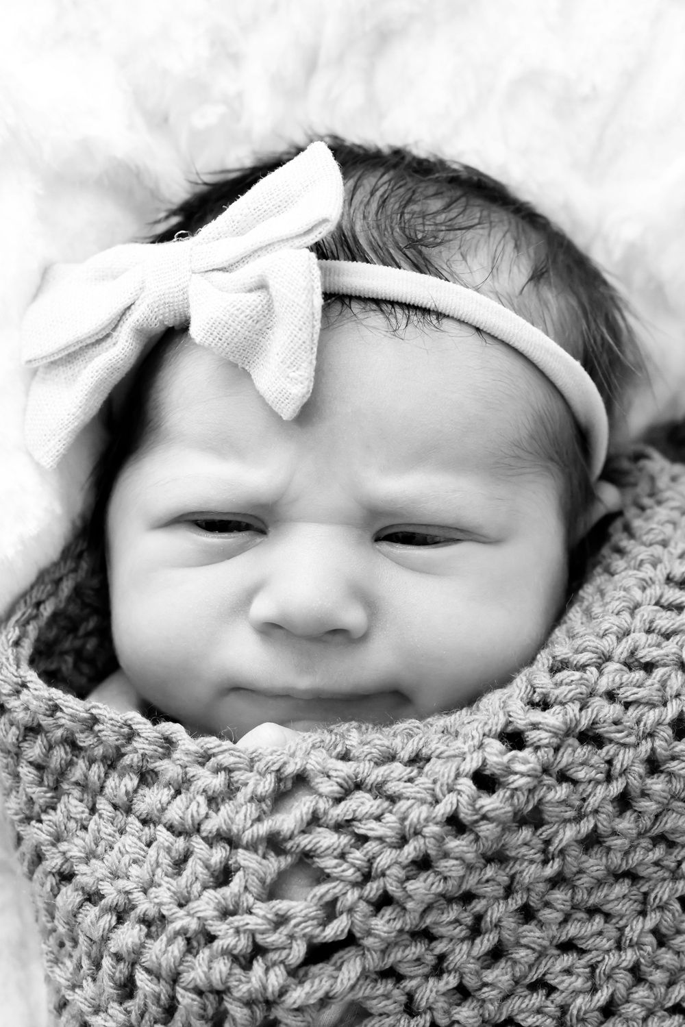 Black and white portrait of newborn baby wrapped in knit blanket wearing a small bow headband.