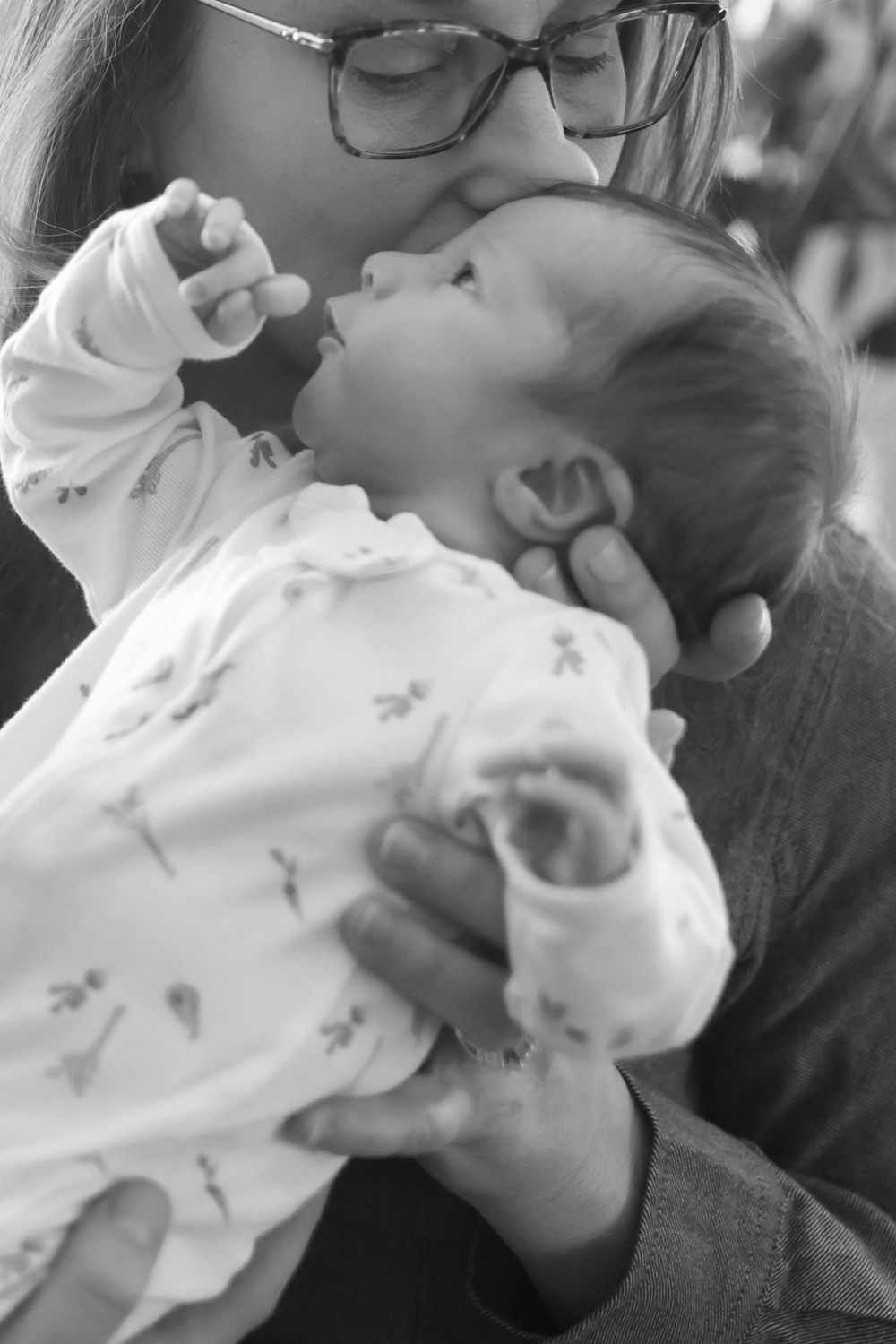 Black and white photo of mother with glasses kissing her alert newborn baby dressed in white onesie.