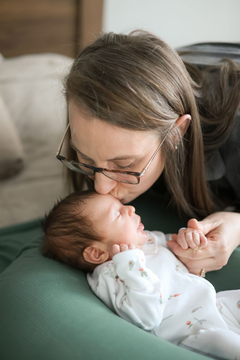 Mother with glasses gently kissing her sleeping newborn baby while holding tiny hand on green bed.