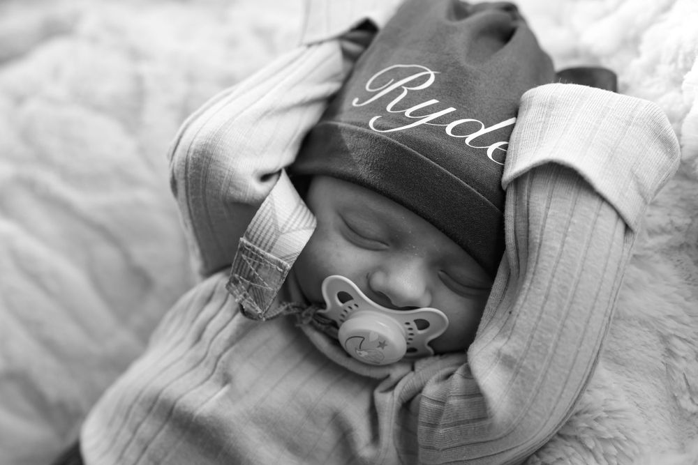 Sleeping newborn baby wearing personalized hat named Ryder with pacifier.