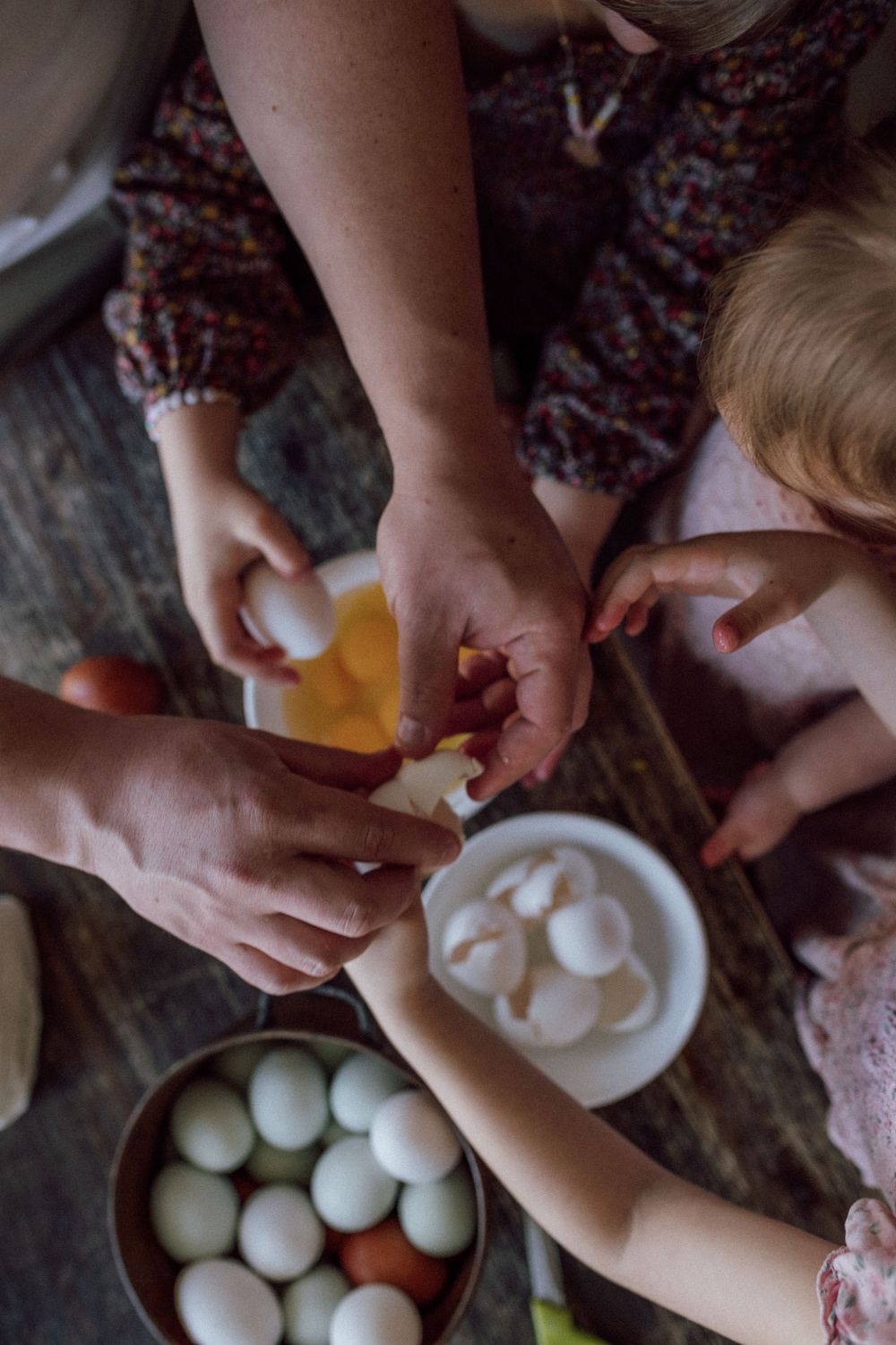 Several hands work together peeling eggs at a wooden table with bowls of fresh eggs nearby. in home family newborn photography session