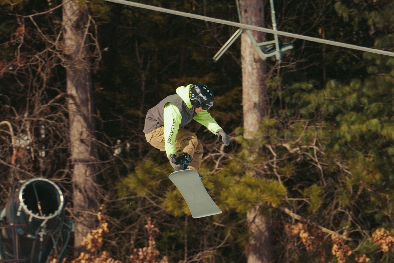 Snowboarder executes mid-air trick above snow-covered slopes with trees in background.
