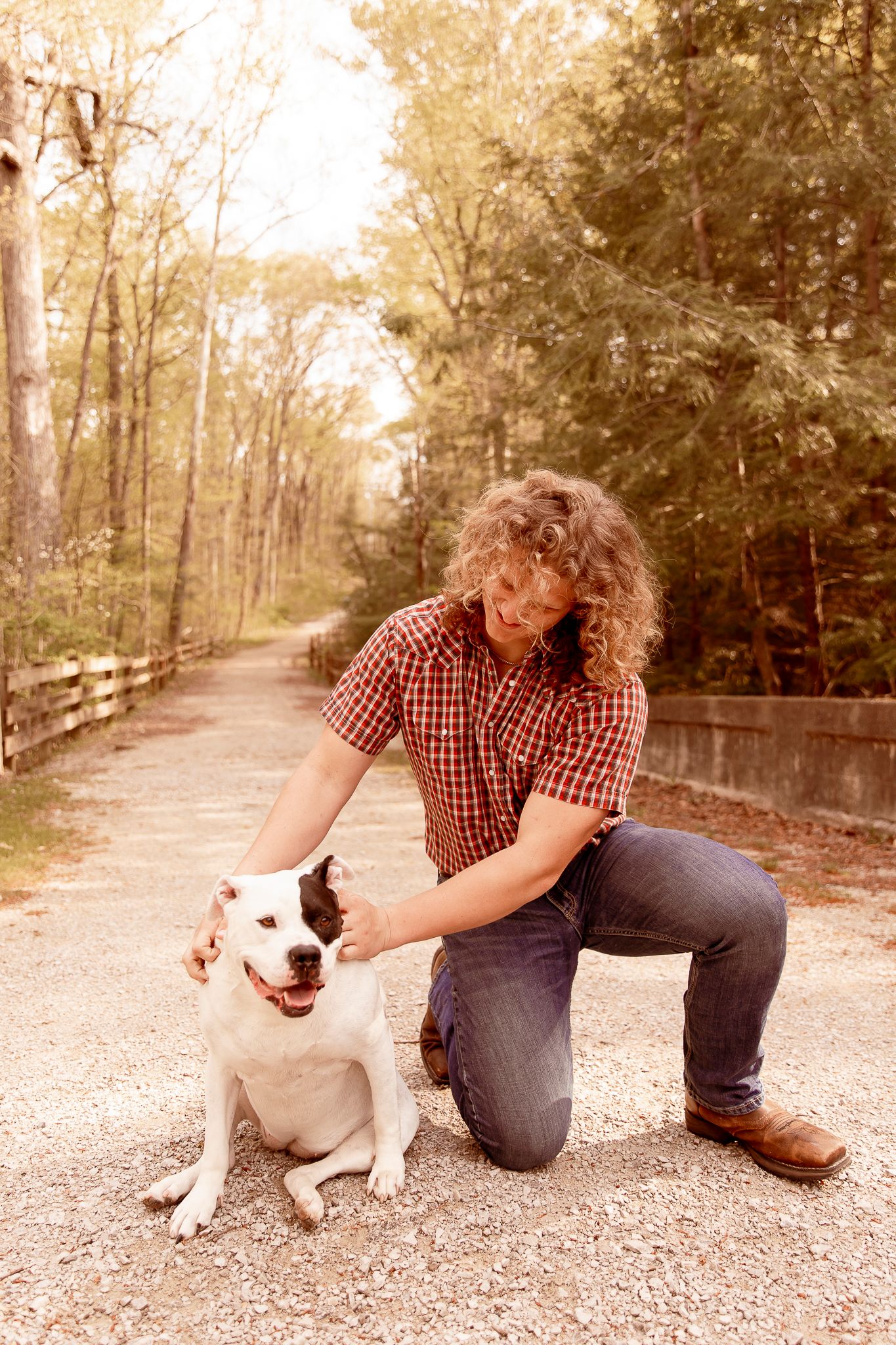 A person in a plaid shirt and jeans kneels beside a happy white and brown spotted dog on a gravel path in the woods.
