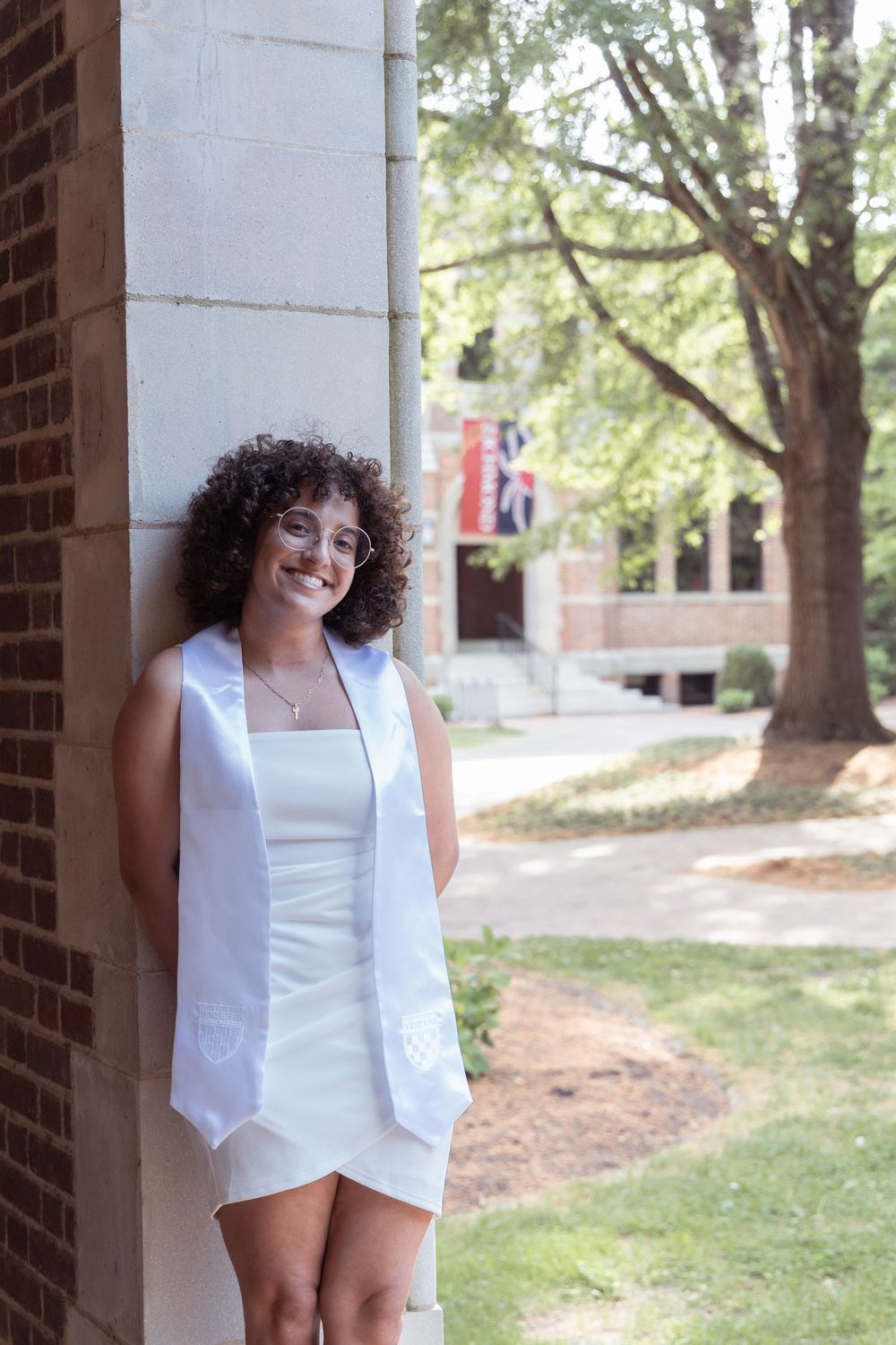 Graduate in white attire poses against brick column during commencement.