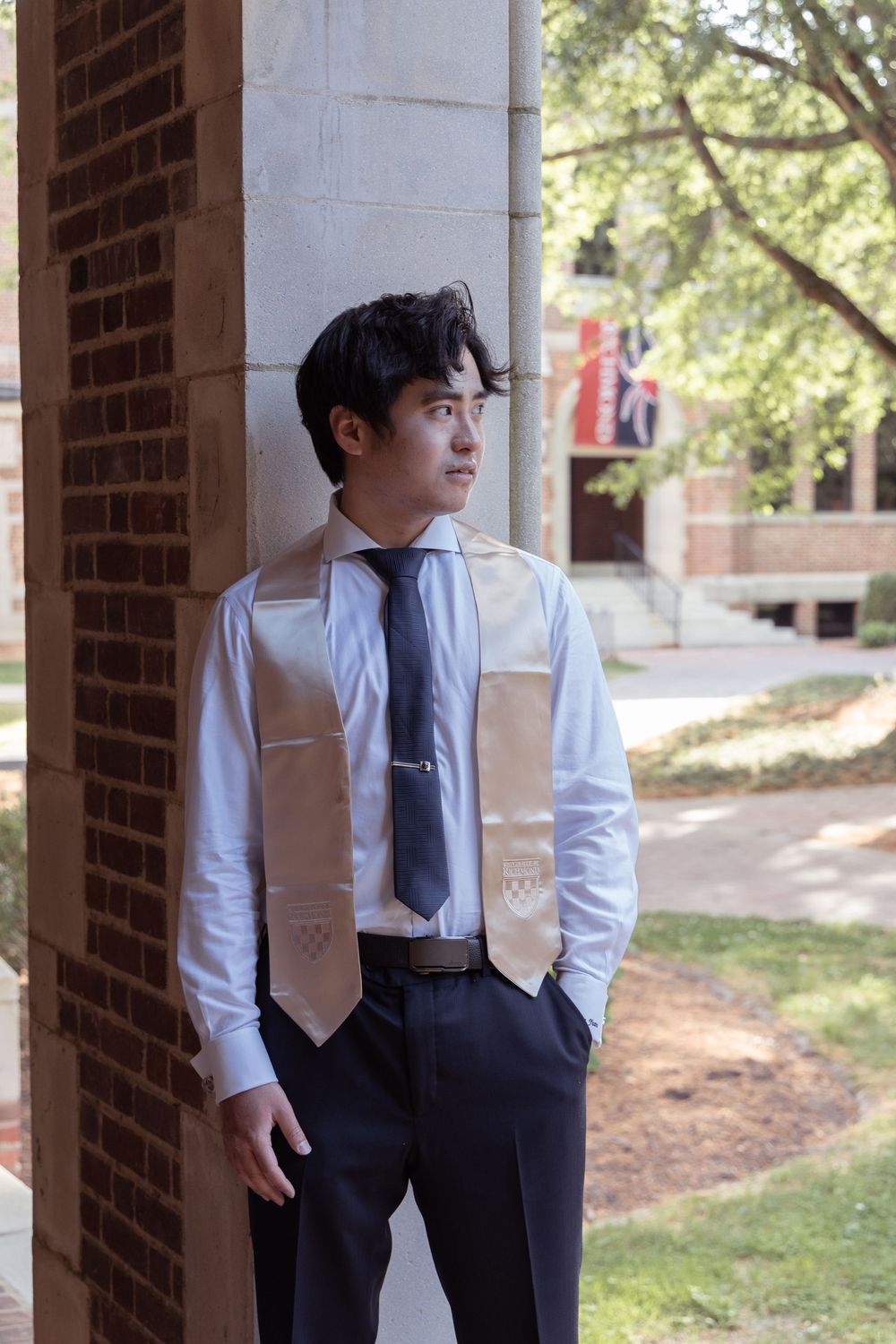 Graduate in formal attire poses against brick column at university campus.