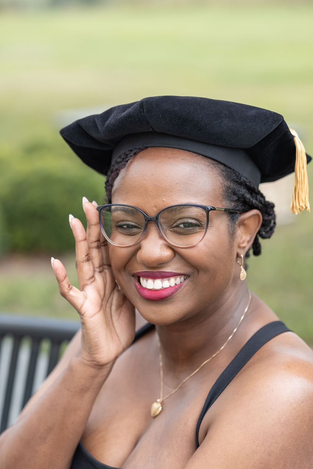 Graduate in academic regalia smiles joyfully while wearing glasses and graduation cap during commencement ceremony.
