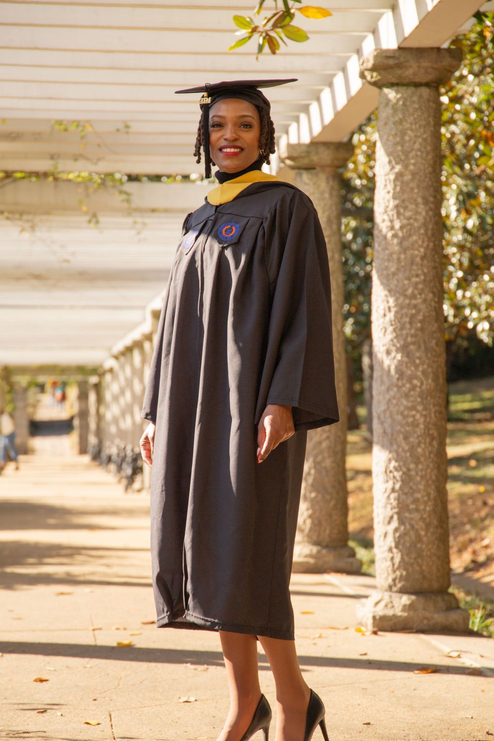 Graduate in traditional black academic regalia poses under white pergola.