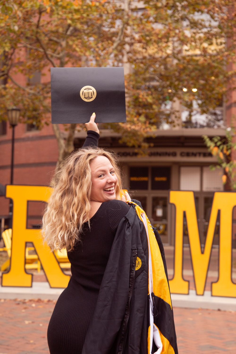 Graduate holding diploma overhead with RAM letters visible in background.
