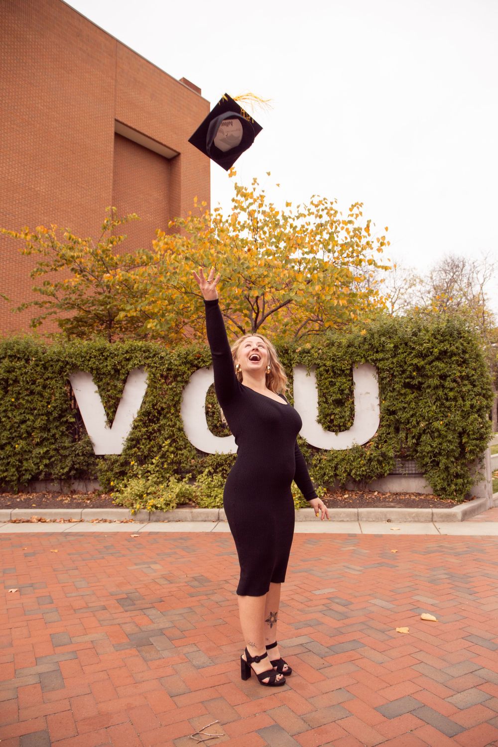 Graduate celebrating by tossing cap in front of VCU letters on brick courtyard.