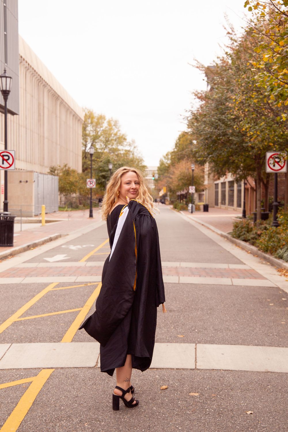 Graduate in academic regalia poses on empty street during autumn.