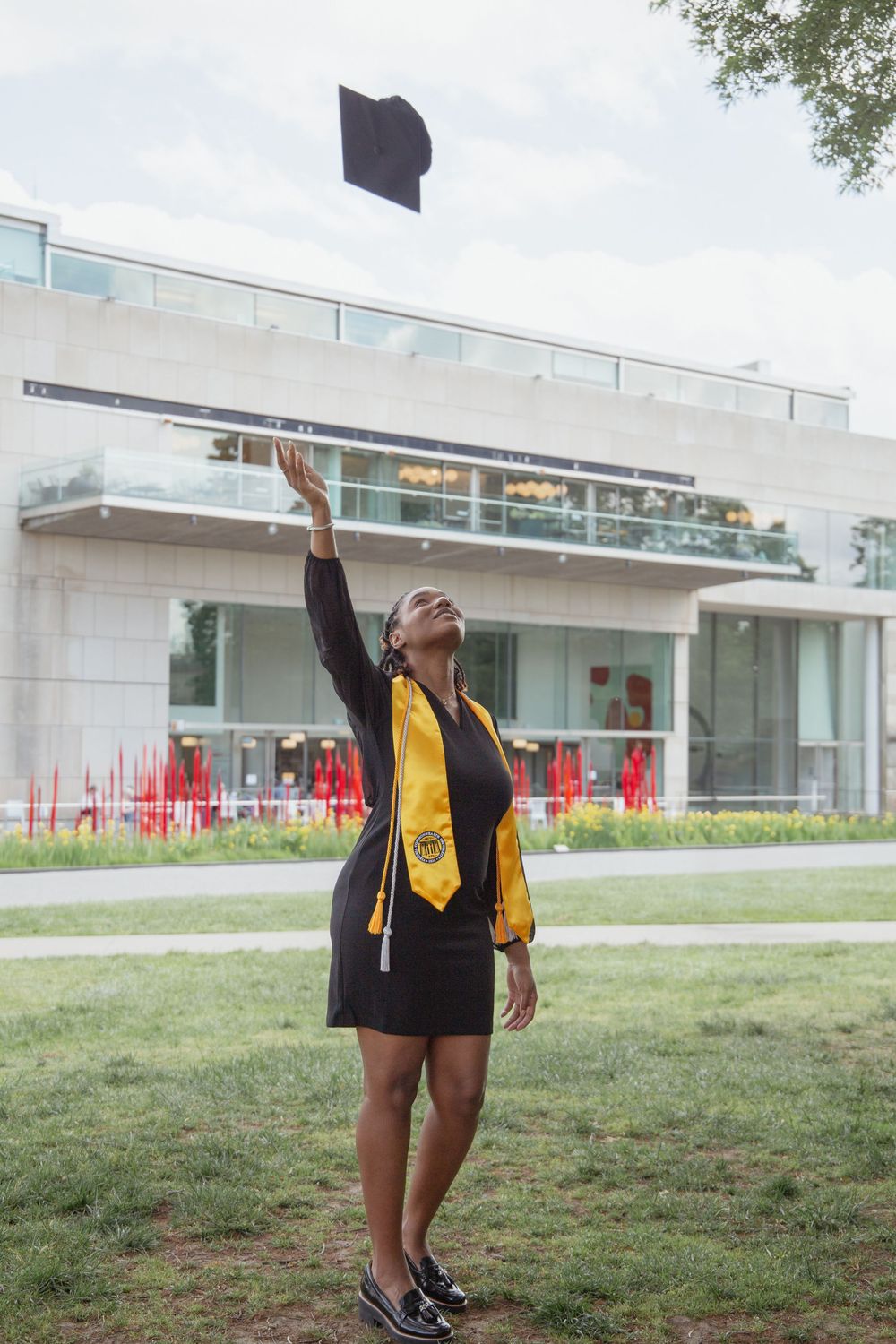 Graduate in black and gold regalia throws cap in celebration on campus lawn.