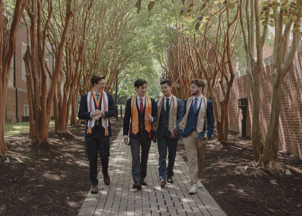 Four graduates walking together along a tree-lined brick path wearing caps and gowns with orange stoles.