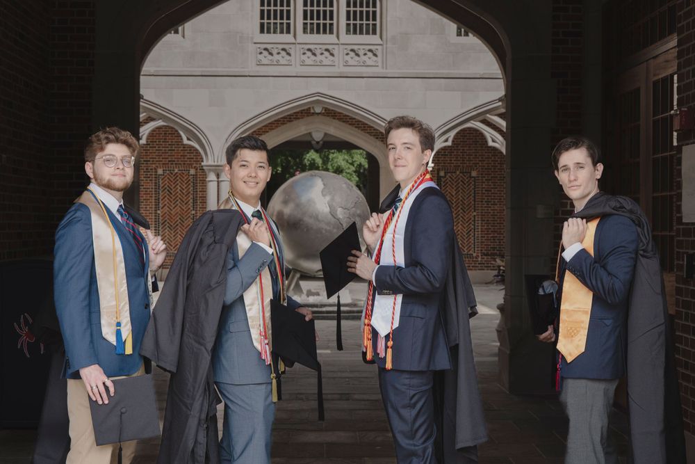 Graduates posing in front of a globe monument on campus wearing academic regalia and orange stoles.