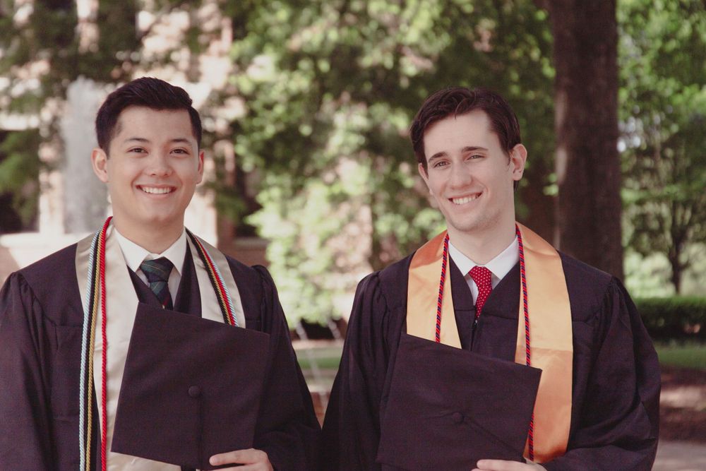 Two graduates standing together outdoors in academic regalia with colorful stoles and smiling.