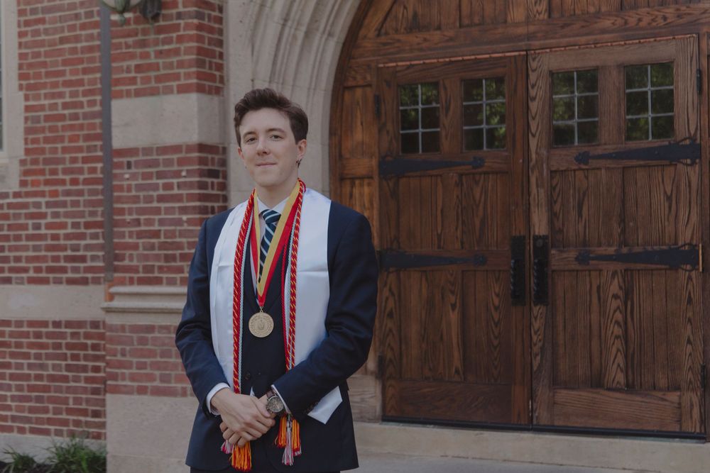 Graduate in academic regalia with stoles and medals standing in front of ornate wooden doors.