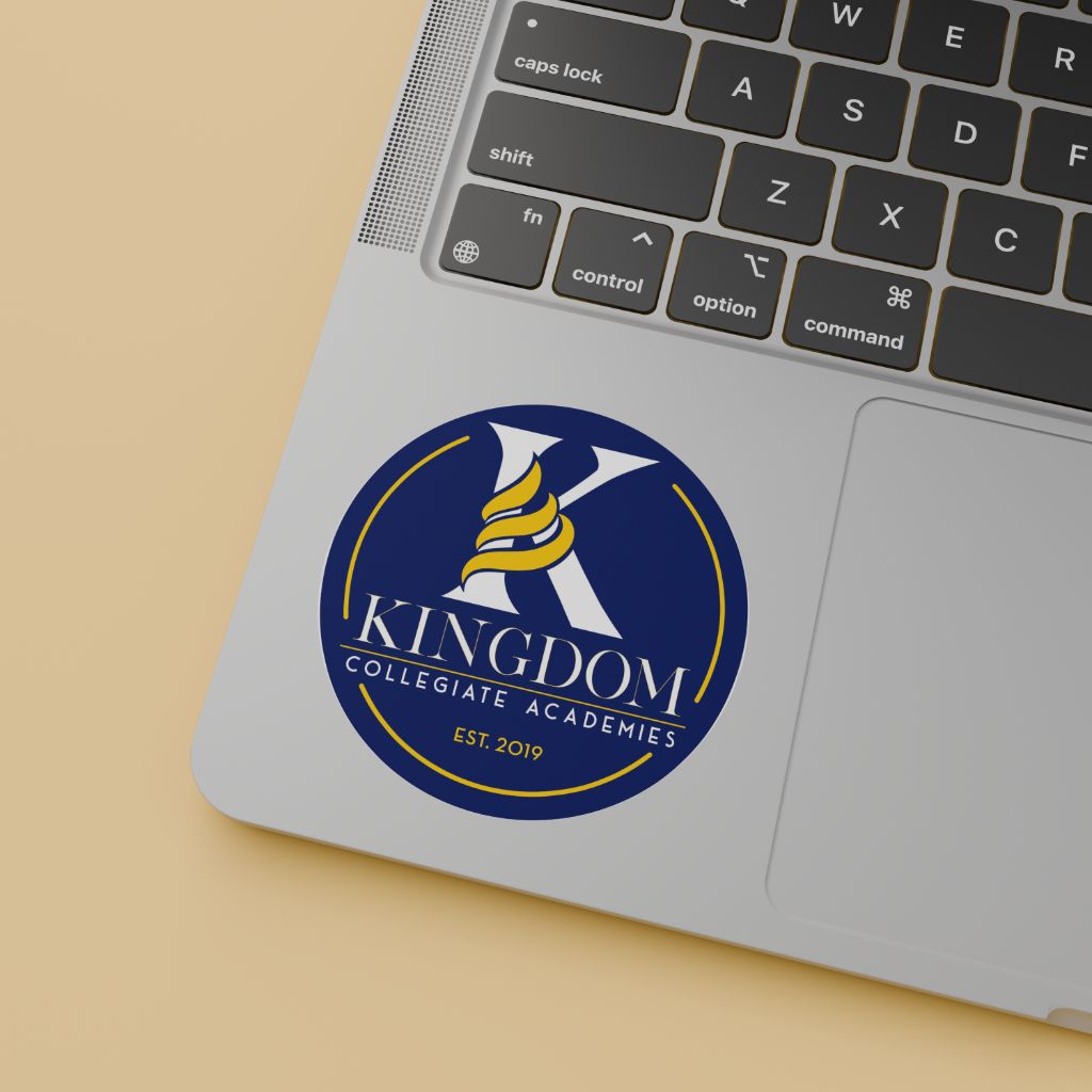 A round blue Kingdom Collegiate Academy logo sticker displayed on a silver laptop keyboard.