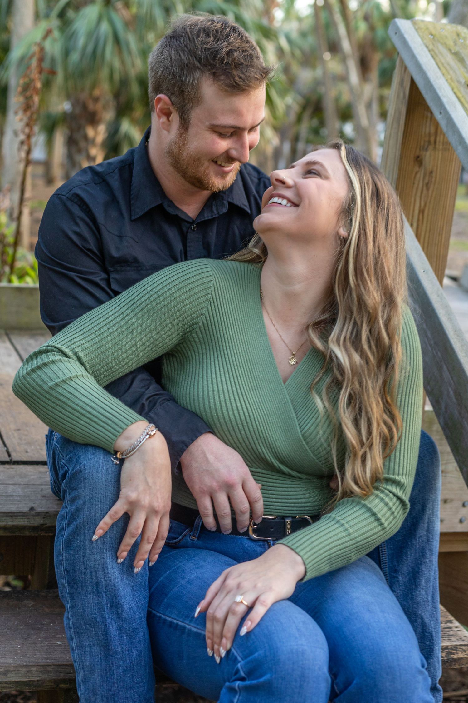 Happy couple laughing together sitting on wooden steps, woman showing engagement ring in green sweater.