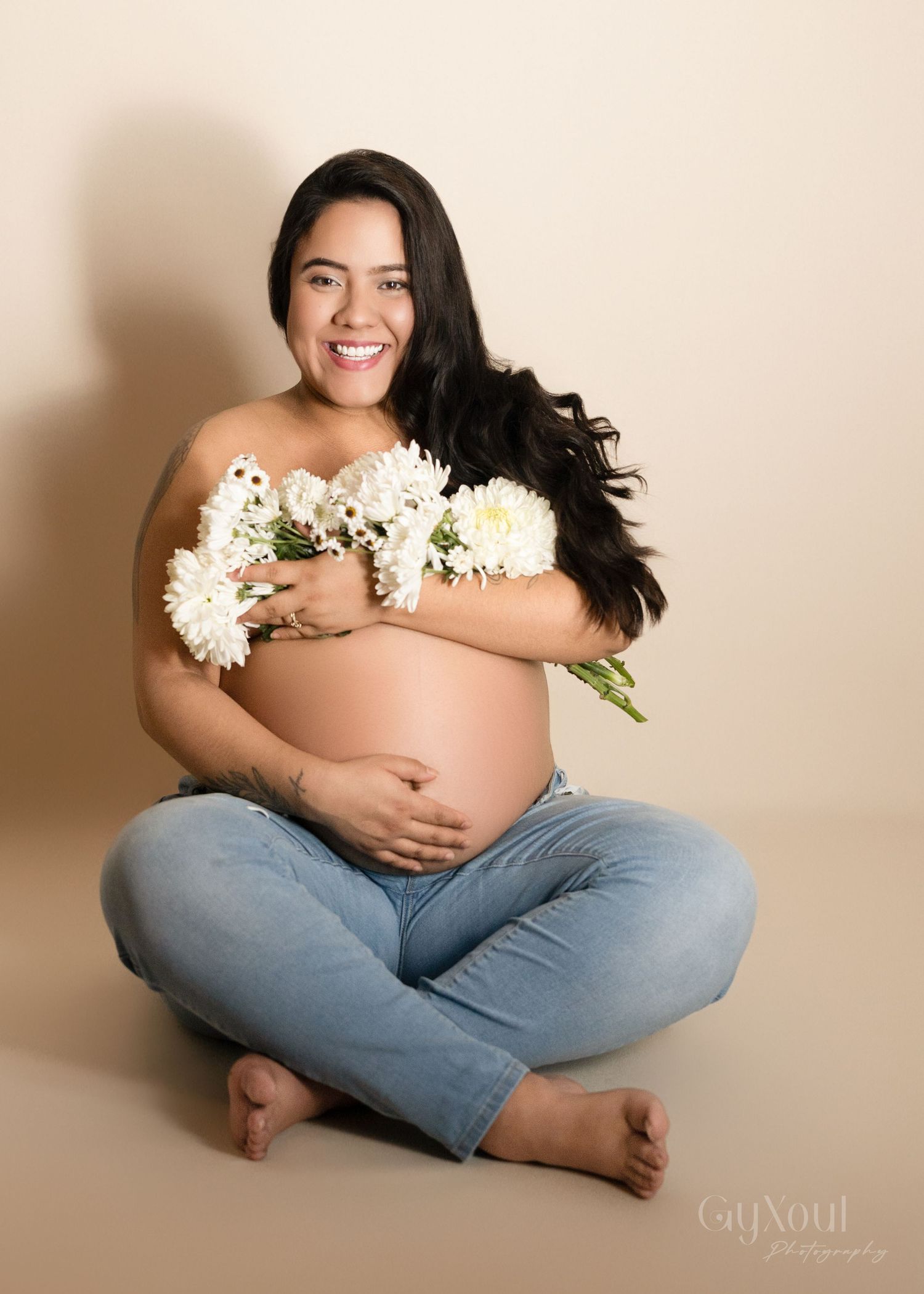 Casual maternity portrait with white flowers and denim jeans against beige background.